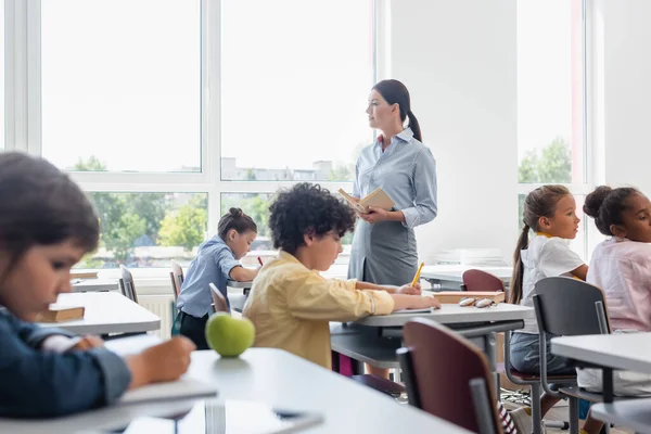 Enfoque Selectivo Del Profesor Sosteniendo Libro Cerca Alumnos Multiculturales Escribiendo — Foto de Stock