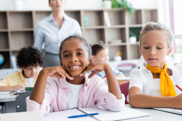 Selective focus of african american schoolgirl looking at camera near classmates and teacher in school 
