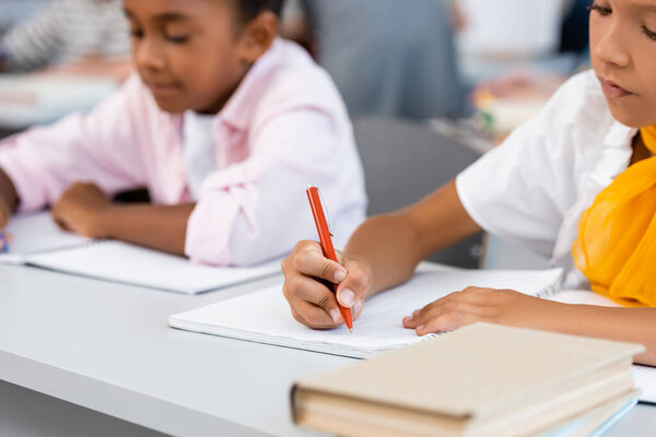 Selective focus of multiethnic schoolgirls writing on notebooks near books on desk in classroom 