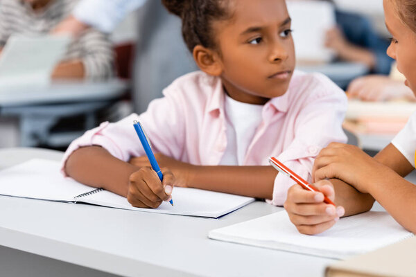Selective focus of schoolgirl talking to african american friend while writing on notebook in classroom 