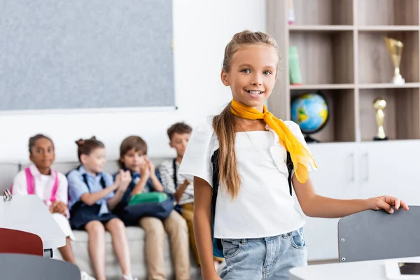 Selective focus of schoolgirl standing near desk with multiethnic classmates at background