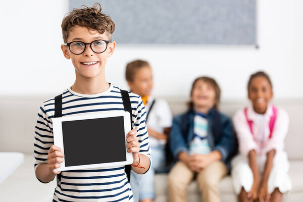 Selective focus of schoolboy in eyeglasses showing digital tablet with blank screen in classroom 