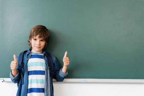 Schoolboy showing thumbs up while standing near green chalkboard 