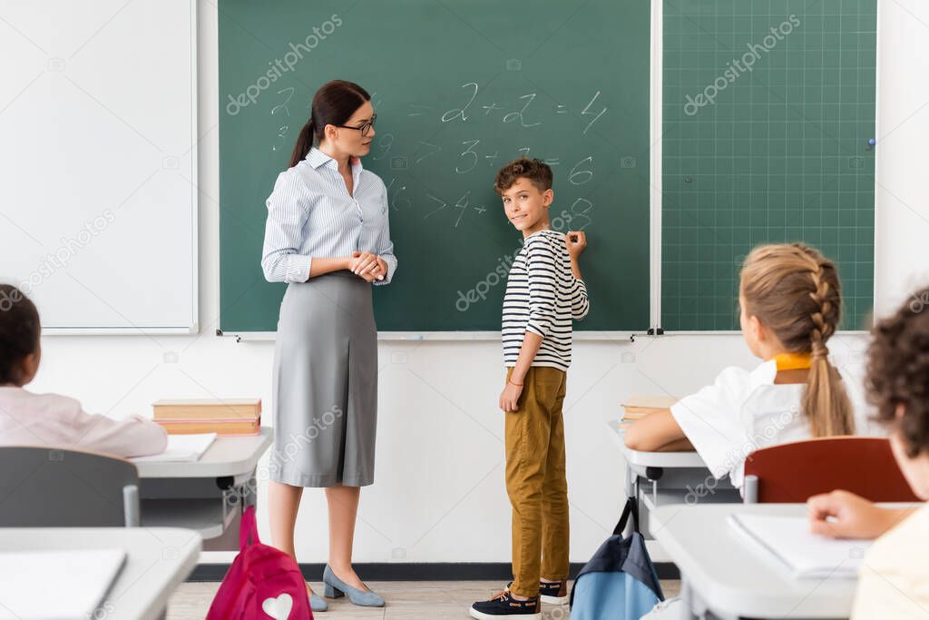 schoolboy looking at multicultural classmates while solving equations on chalkboard during math lesson
