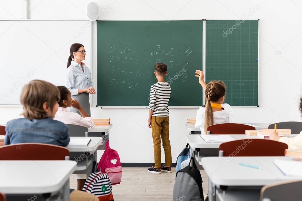 back view of schoolboy solving equations on chalkboard near teacher and multicultural classmates