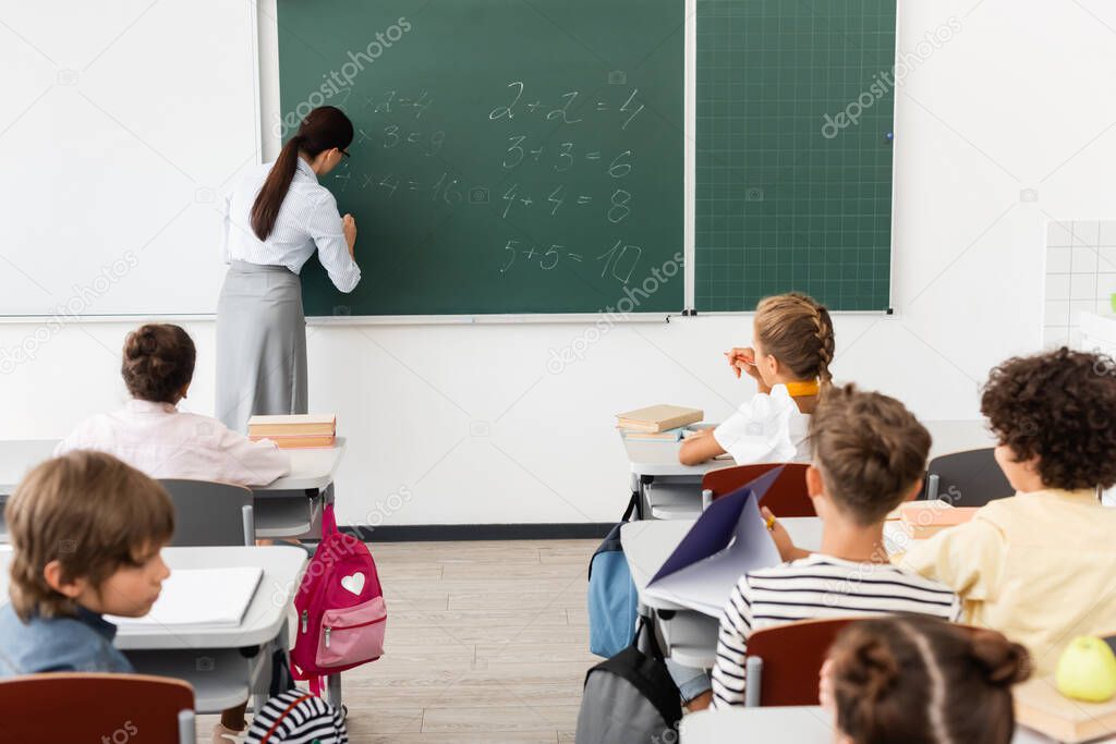 back view of teacher writing equations on chalkboard near multicultural pupils during math lesson