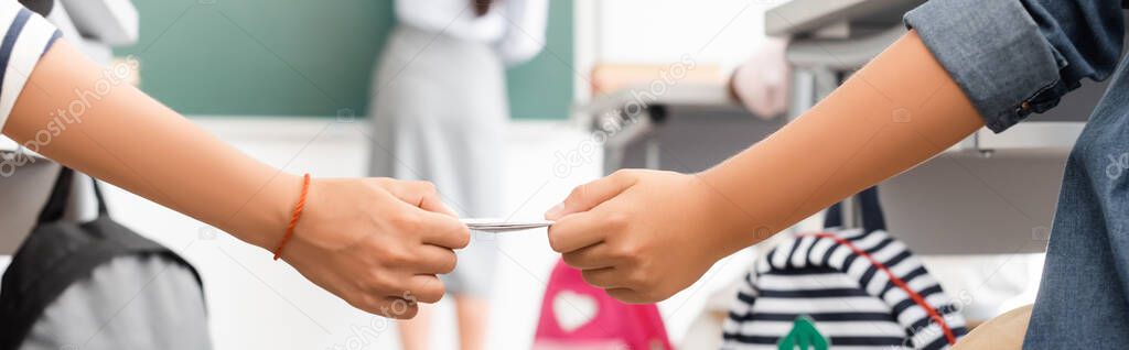 cropped view of schoolboy passing note to classmate while teacher standing at chalkboard, horizontal image