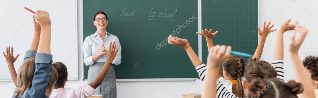 back view of multicultural pupils with hands in air, and teacher standing at chalkboard with back to school inscription, horizontal image