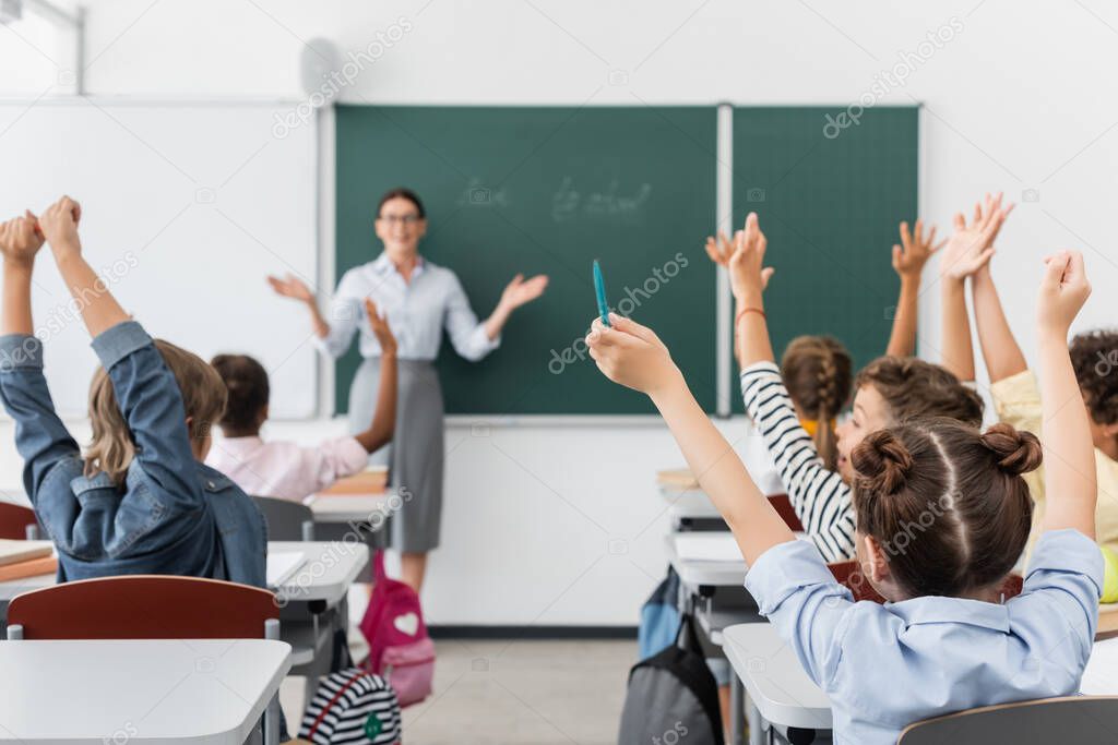 back view of multicultural pupils with hands in air, and teacher standing at chalkboard with open arms