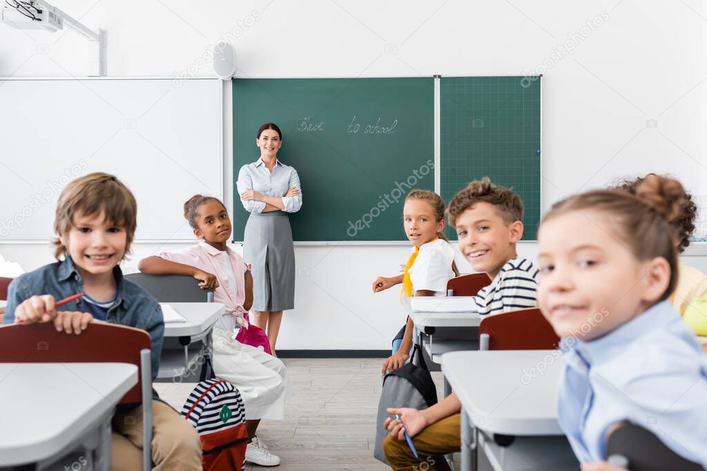 teacher with crossed arms and multicultural pupils looking at camera during lesson