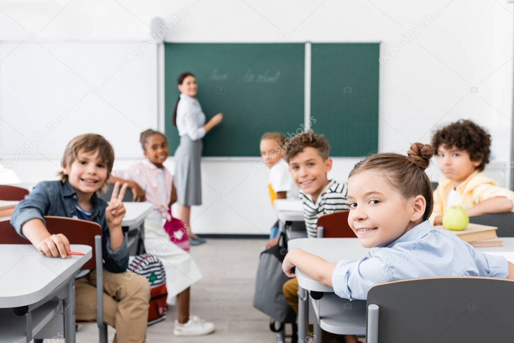 selective focus of multicultural pupils looking at camera, and teacher standing at chalkboard on background
