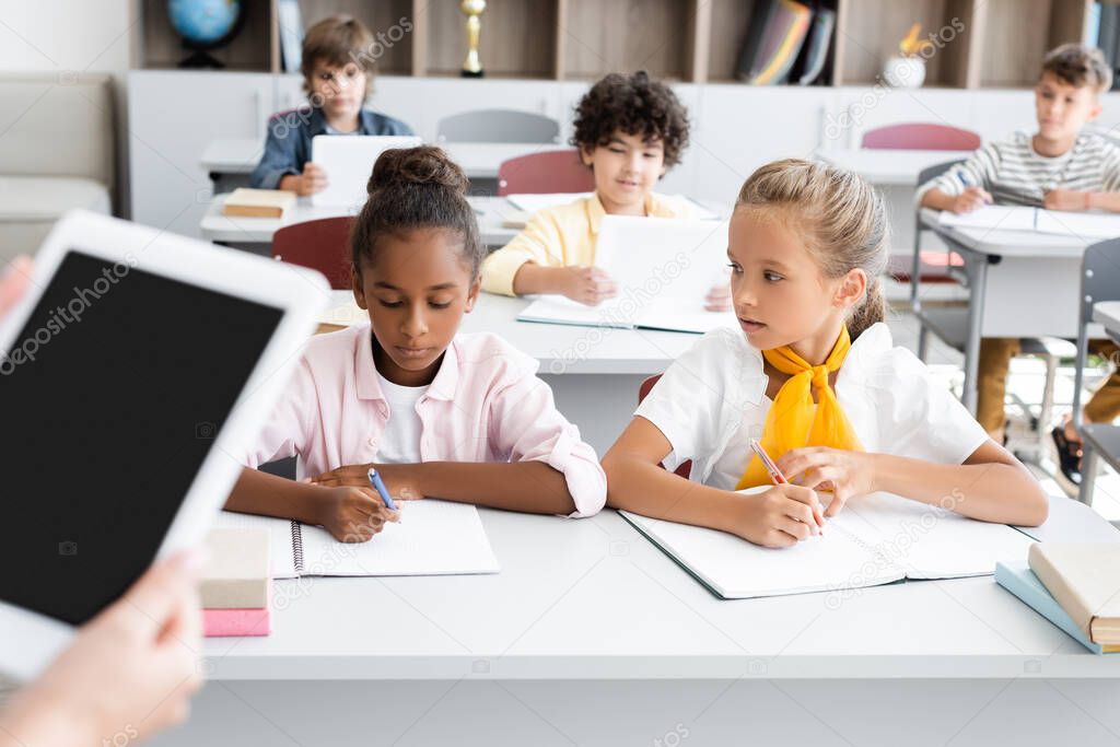 cropped view of teacher holding digital tablet with blank screen near multicultural pupils writing in notebooks