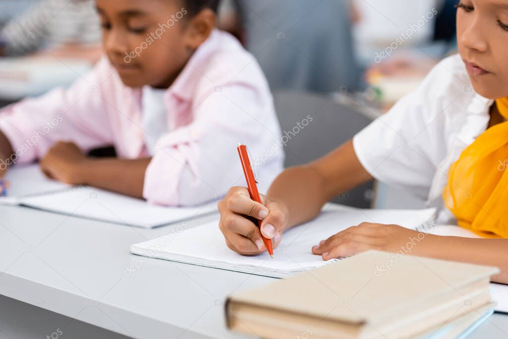 Selective focus of multiethnic schoolgirls writing on notebooks near books on desk in classroom 
