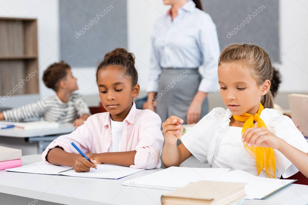 Selective focus of multcultural schoolgirls sitting near books and notebooks on desk in classroom 