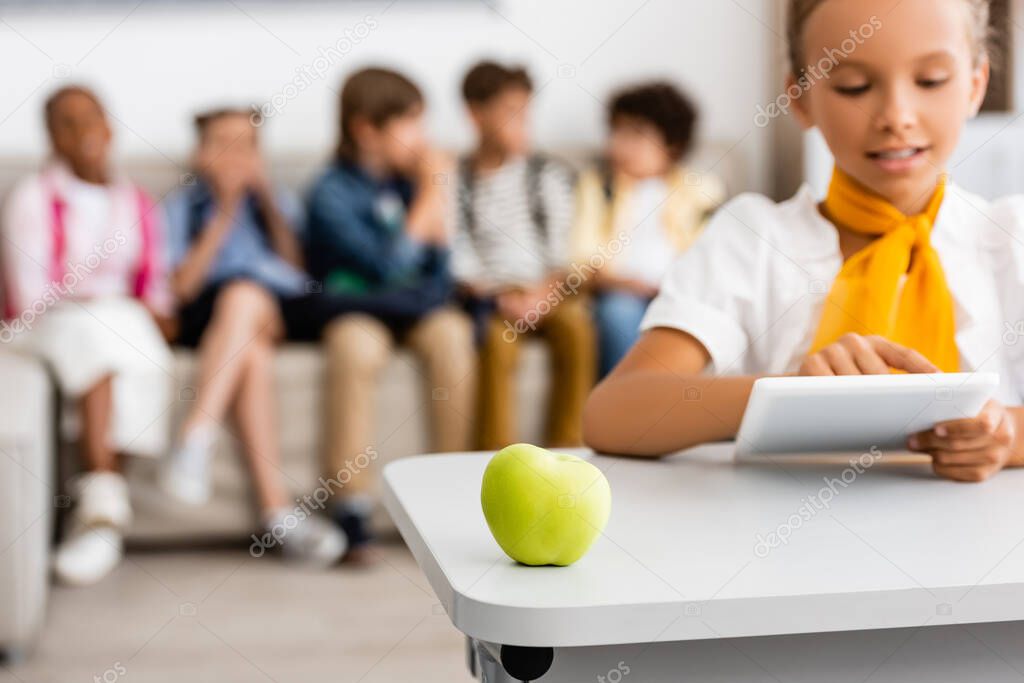 Selective focus of fresh apple on desk near schoolgirl using digital tablet in classroom 