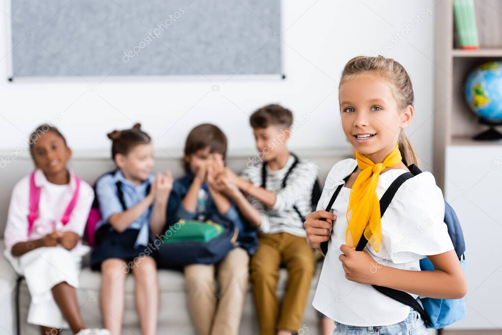 Selective focus of schoolgirl with backpack looking at camera with multiethnic friends at background in school 