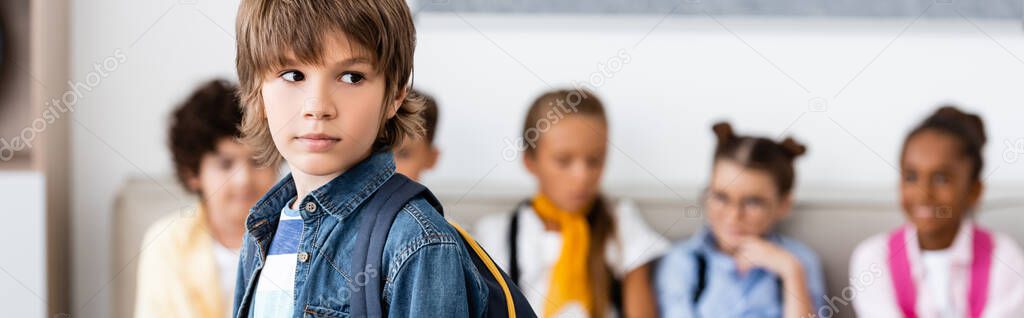Panoramic image of schoolboy with backpack standing near multiethnic classmates in school 