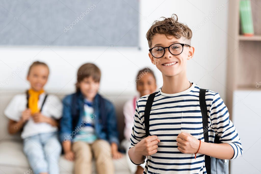 Selective focus of schoolboy with backpack and eyeglasses looking at camera with classmates at background 