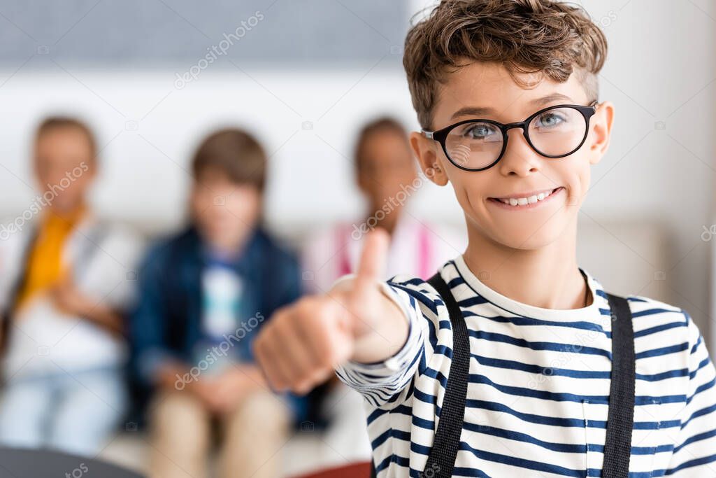 Selective focus of schoolboy in eyeglasses showing thumb up at camera in classroom 