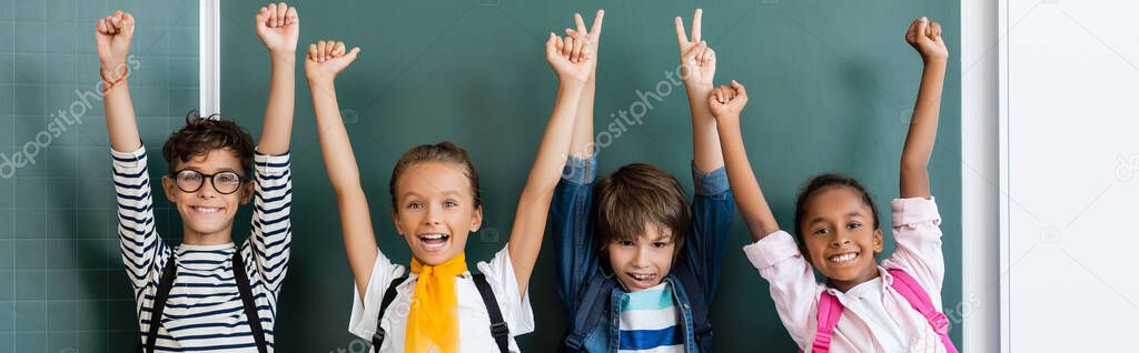 Horizontal crop of multiethic classmates with backpacks showing yeah and peace gestures near chalkboard 