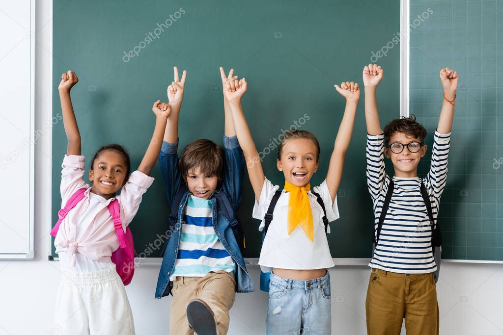 Selective focus of multicultural classmates showing peace and yes gestures near chalkboard in classroom 