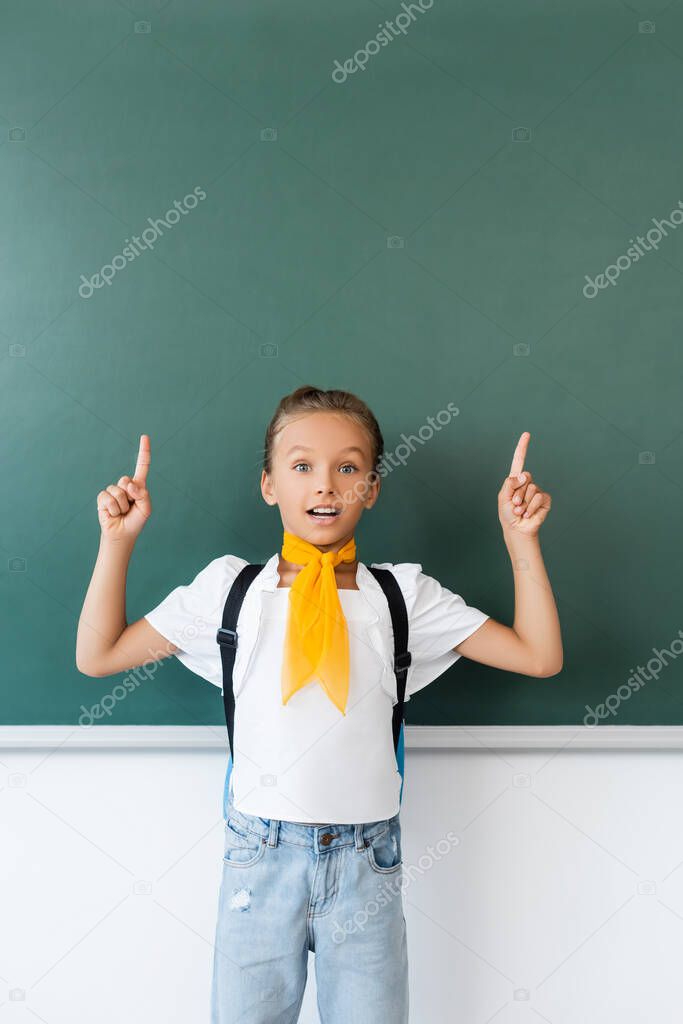 Excited schoolgirl with backpack pointing with fingers at chalkboard 