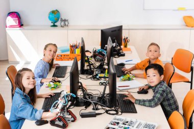 Selective focus of multicultural schoolkids looking at camera near robots and computers in classroom  clipart
