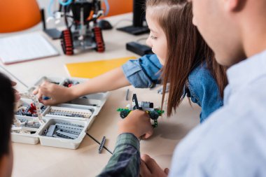 Selective focus of teacher sitting near schoolkids modeling robot in classroom  clipart