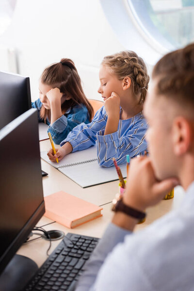 Selective focus of schoolgirls writing on notebook near teacher and computers in classroom 