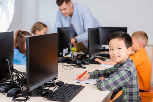 Selective focus of asian schoolboy looking at camera while writing on notebook near computers in stem school 