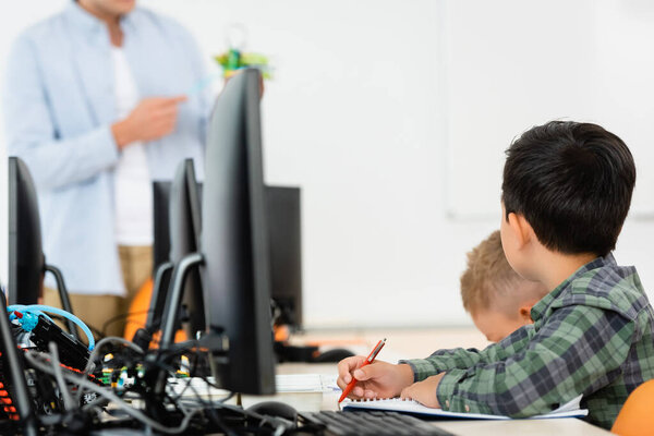 Selective focus of multiethnic schoolboys sitting near computers during lesson with teacher in stem school 