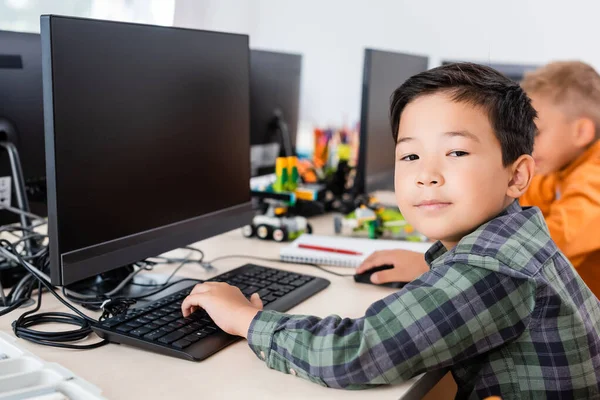 教室でコンピュータを使いながらカメラを見るアジアの少年の選択的焦点 — ストック写真