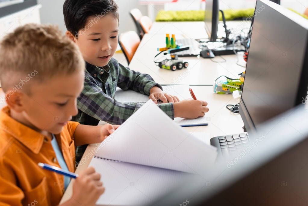 Selective focus of multiethnic schoolboys with notebooks sitting near computers in stem school 