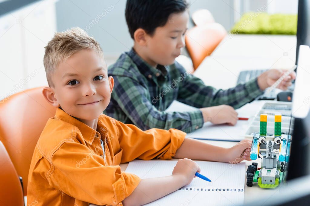 Selective focus of schoolboy looking at camera near robot and asian friend in stem school 