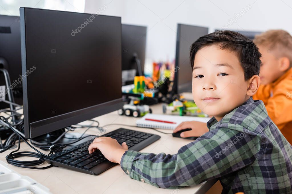 Selective focus of asian schoolboy looking at camera while using computer in classroom 