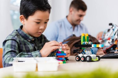 Selective focus of asian schoolboy with building blocks modeling robot near friend and teacher in school  clipart