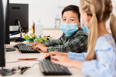 Tıp maskeli Asyalı öğrencinin sınıftaki arkadaşının yanındaki bilgisayarı kullanarak seçici odak noktası. 