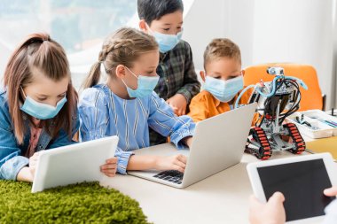 Tıp maskeli çocukların seçici odak noktası okulda öğretmen ve robotun yanında dijital tablet ve dizüstü bilgisayar kullanmaları. 