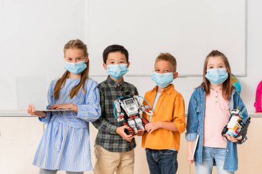 Tıp maskeli çok ırklı okul çocukları kök okulda robot ve dizüstü bilgisayar taşıyorlar. 