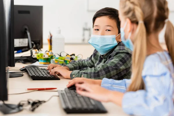 教室で友人の近くのコンピュータを使用して医療用マスクでアジアの学童の選択的焦点 — ストック写真