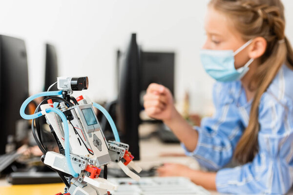 Selective focus of robot on table near schoolgirl in medical mask in classroom 