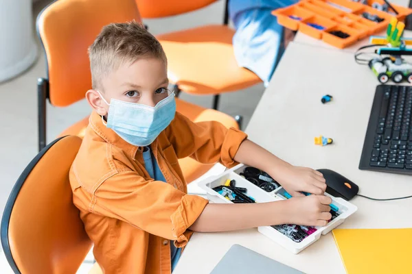 教室のコンピュータの近くに建物のブロックを保持する医療用マスクの学校の少年 — ストック写真