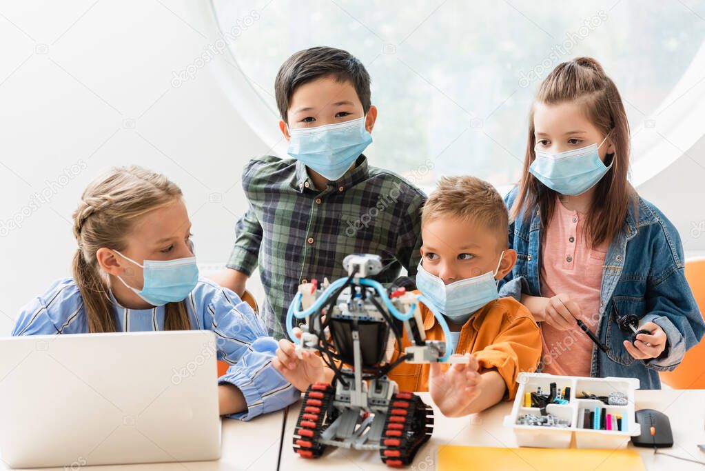 Selective focus of multiethnic classmates in medical masks constructing robot near laptop in classroom 