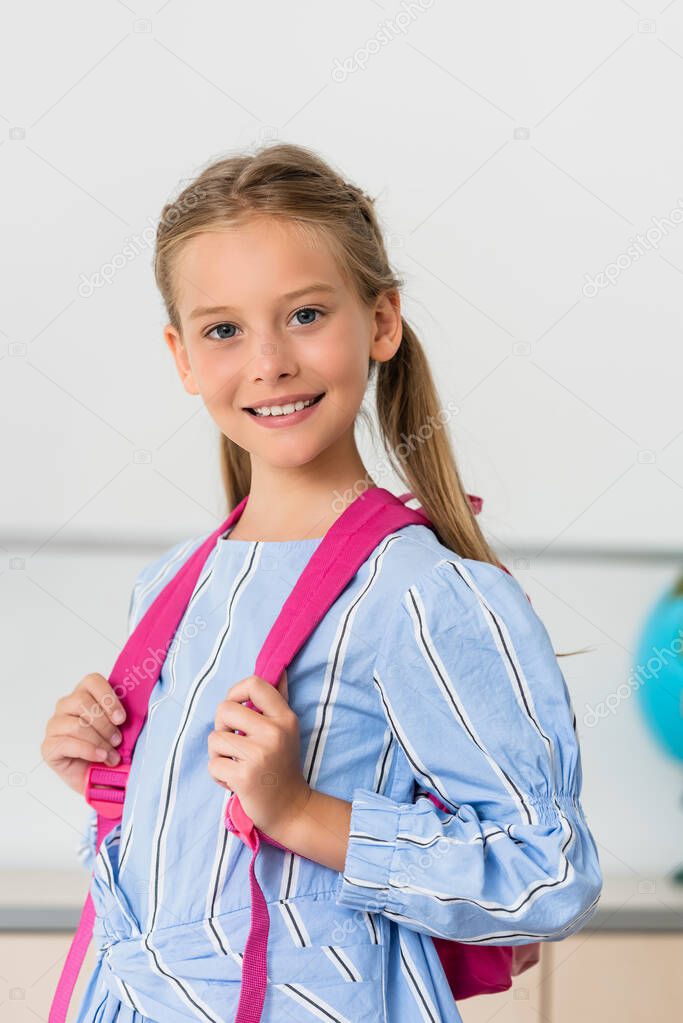 Schoolgirl looking at camera while holding backpack in classroom 