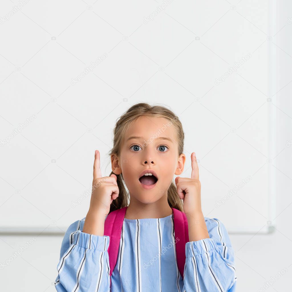 Excited kid with backpack pointing with fingers while looking at camera in school