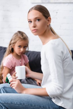Selective focus of young woman holding cup and blisters with pills near ill daughter at home  clipart