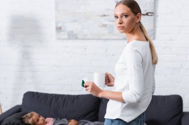 Selective focus of young woman with mug and pills looking at camera near sick child on couch at home  clipart