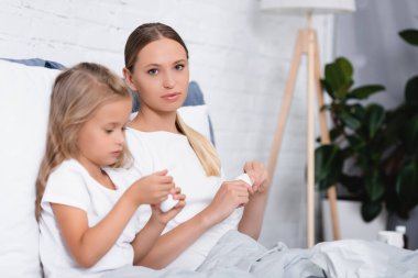 Selective focus of young woman holding jar with pills and looking at daughter on bed  clipart
