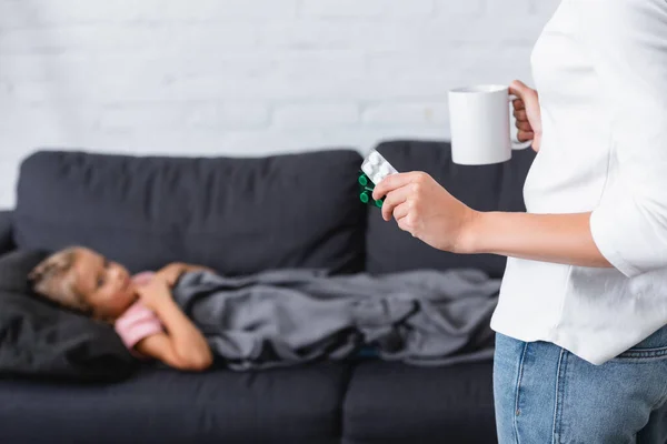 Selective focus of woman with cup and pills standing near sick daughter on couch