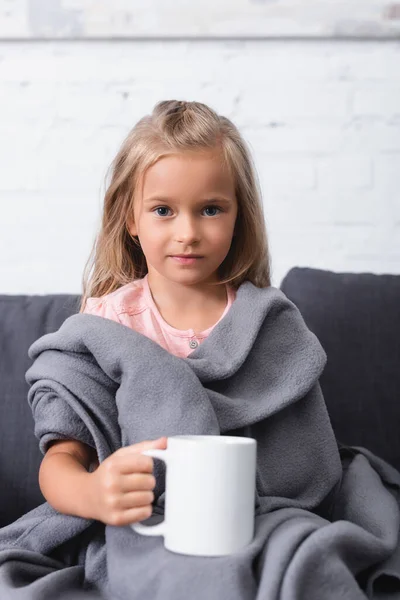 ソファにカップを保持しながら 子供の選択的な焦点は カメラを見て毛布に包まれた — ストック写真